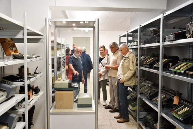 In zwei gegenüberstehenden Regalen im Schaudepot stehen Rechen- und andere Maschinen aus BWS-Produktion. In der Mitte des Ganges zwischen den Regalen steht eine Glasvitrine im Hochformat. Darin wird eine Rechenmaschine präsentiert. Um die Vitrine herum steht steht eine Gruppe von Menschen. Einer der Ehrenamtlichen des Schaudepots erklärt anhand der Rechenmaschine in der Glasvitrine deren Funktionsweise.