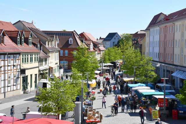 Blick von oben auf eine längere Einkaufsstraße mit Häusern rechts und links sowie davor stehenden Marktsständen, zwischen denen Leute flanieren.
