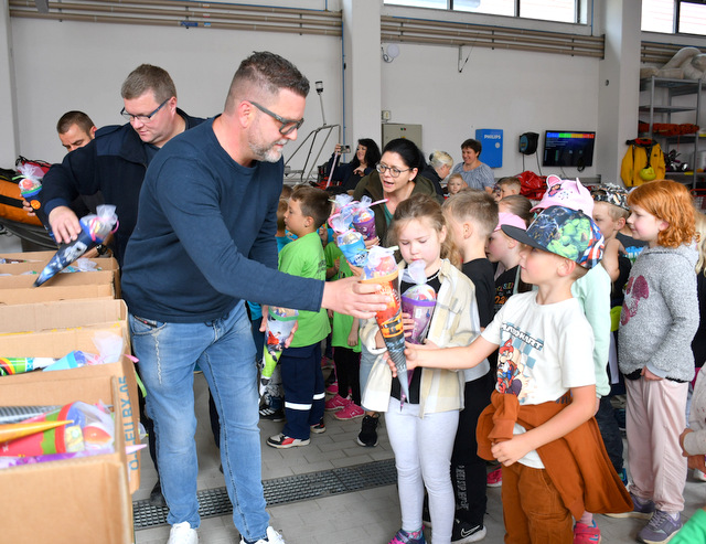 Viele Kinder stehen in einer großen Halle vor drei Tischen mit gefüllten mittelgroßen Zuckertüten. Diese werden ihnen von drei Männern überreicht.