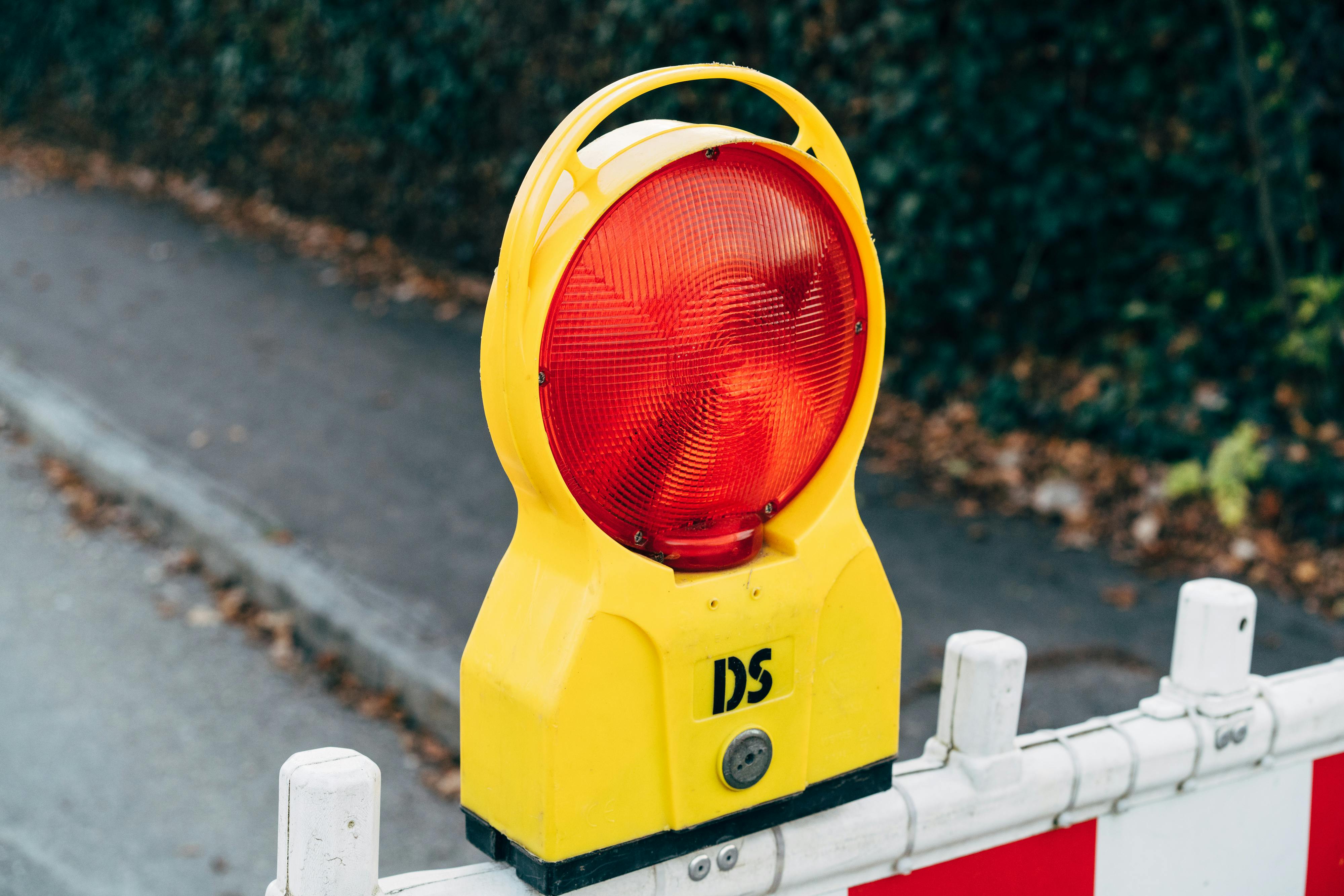 Gelbe Baustellenwarnleuchte mit rotem Blinklicht auf einer weiß-roten Absperrung am Straßenrand. Im Hintergrund ist eine asphaltierte Straße und ein Gehweg mit Laub zu sehen.